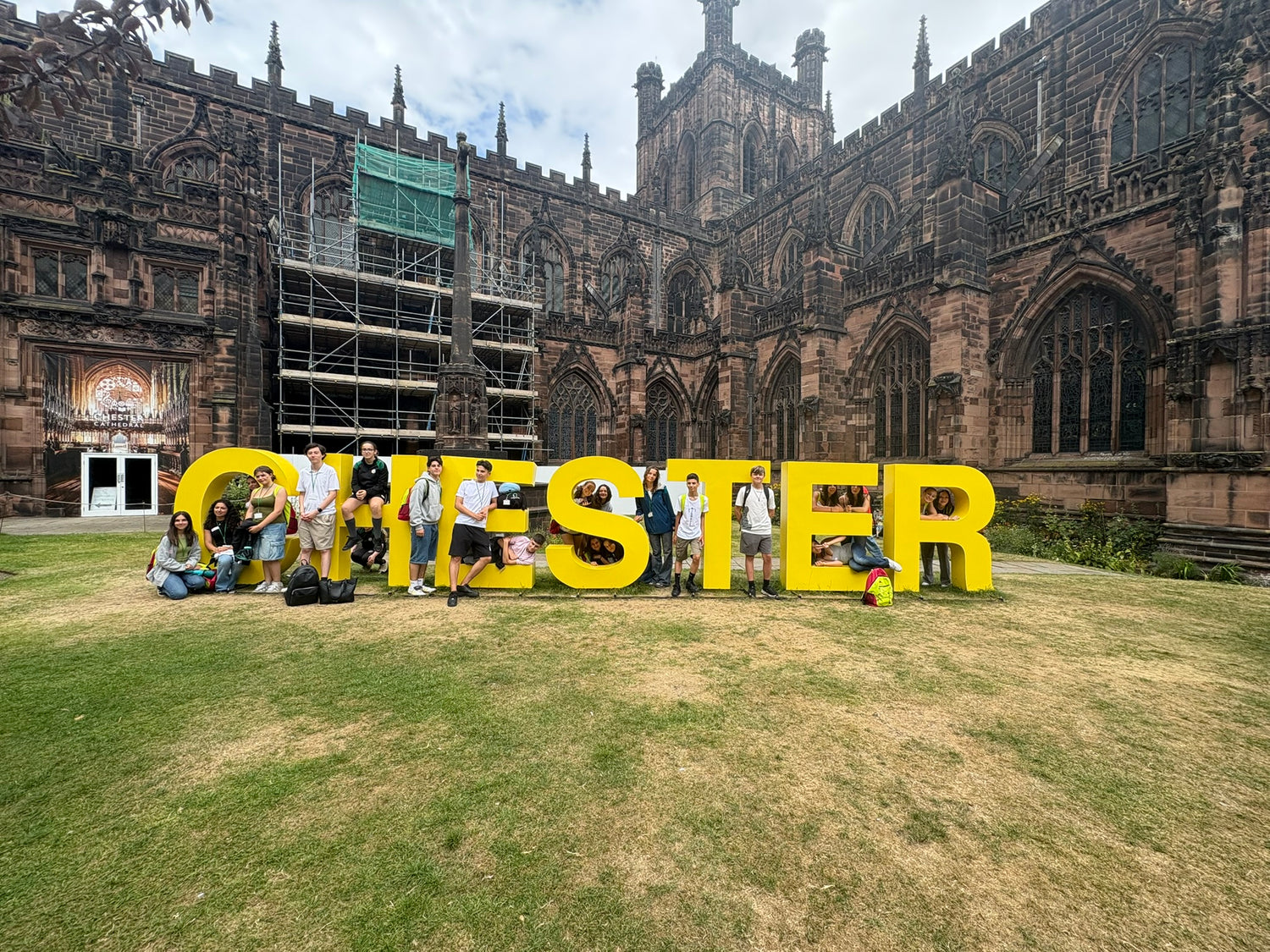 Group of people posing with large yellow letters spelling 'CHESTER' in front of a cathedral.