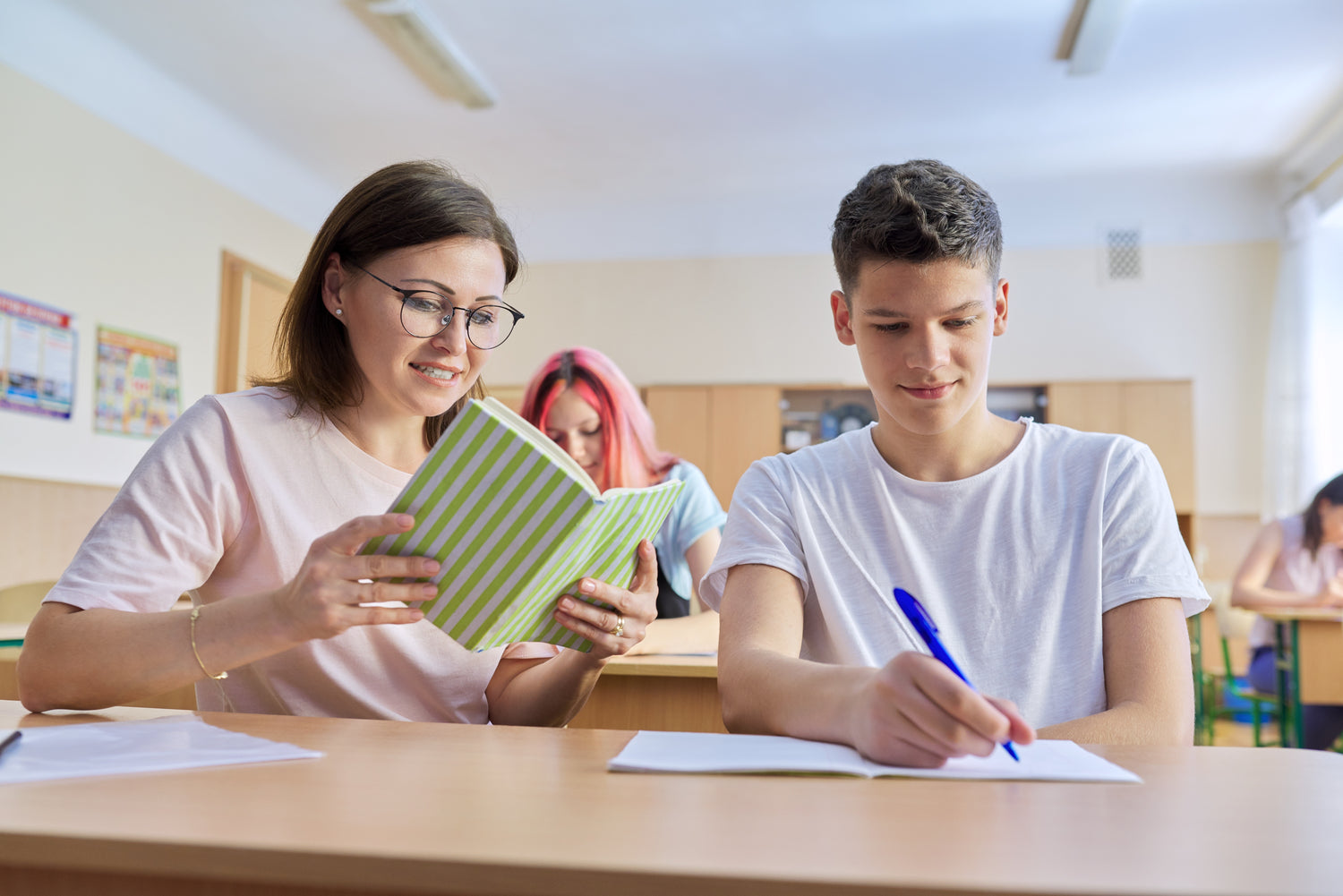 A student and a Teacher are sitting at a desk in a classroom, one holding a notebook and the other writing.